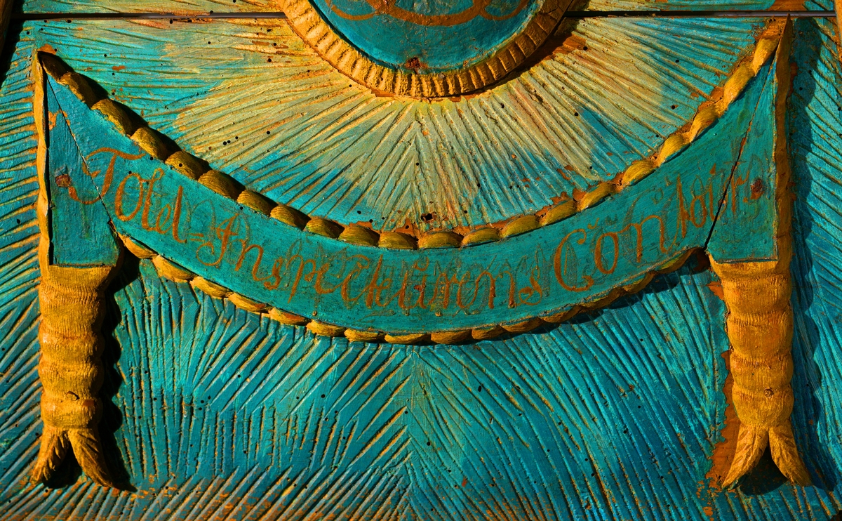 Døroverstykke bestående av 3 sammensatte trebord med not og fjær (bunnplaten). Det har en trekantet hovedform med rett underkant, innadbuede sider og utadbuet overkant. Bunnplaten er utskåret og i flere nyanser av blått, påsatt 2 forhøyninger. Den øverste er oval, blåmalt med krone i gull og rødt over "CR" påmalt i brunt, og langs kanten en lysutskåret bord. Den nederste er formet som en girlander og er avlang, buet og blåmalt med "Told-inspekteurens  Contoir" påmalt med brunfarge. Langs kanten er det en lysutskåret bord og på hver side en lys, utskåret dusk. Skiltet omgitt av en rett list i underkant samt en rød og grå buet, profilert list på sidene og i overkant. Disse er malt i noe som minner om en marmoreringsteknikk (bemalt imitasjon av marmor).Tilhører et sett med tre døroverstykker .Jfr. B-bok nr. 518,519,520. Nr. 518 er borte (ToldCassererens Contoir") Antatt fra Christiania iht. B-bok