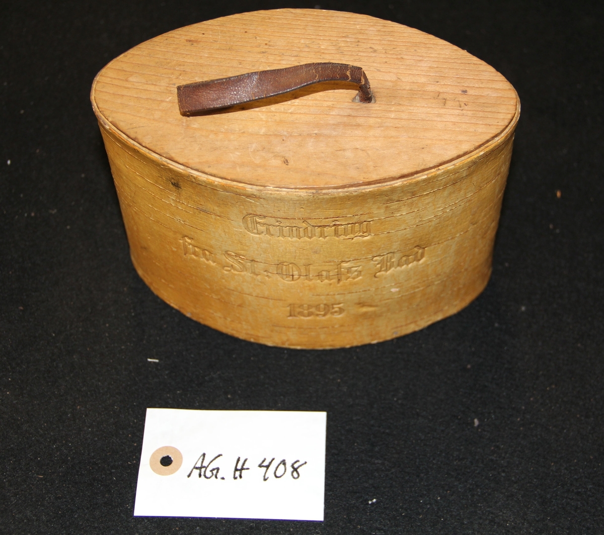 Oval tretine  og lokk med lærhåndtak. Inskribsjon på framsiden av tinen