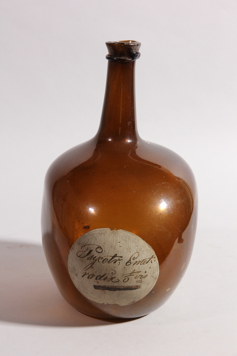 Flaska av brunt glas, rund med tillplattade sidor och hög hals.