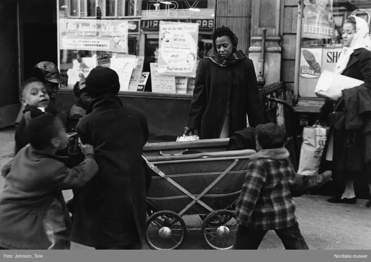 En grupp barn och kvinnor bredvid en barnvagn där en utslagen man ligger. Lenox Avenue, Harlem, New York, USA.