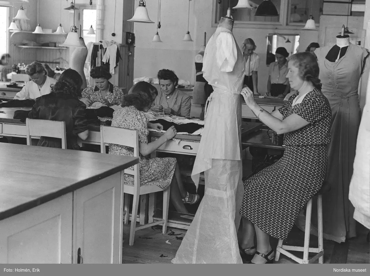 Interiör från NK, NK:s Franska damskrädderi, ateljén. Runt ett stort bord sitter sömmerskor, några kvinnor syr, en uppsätterska modellerar en toall på en provdocka (mannequin). 26 augusti 1943.