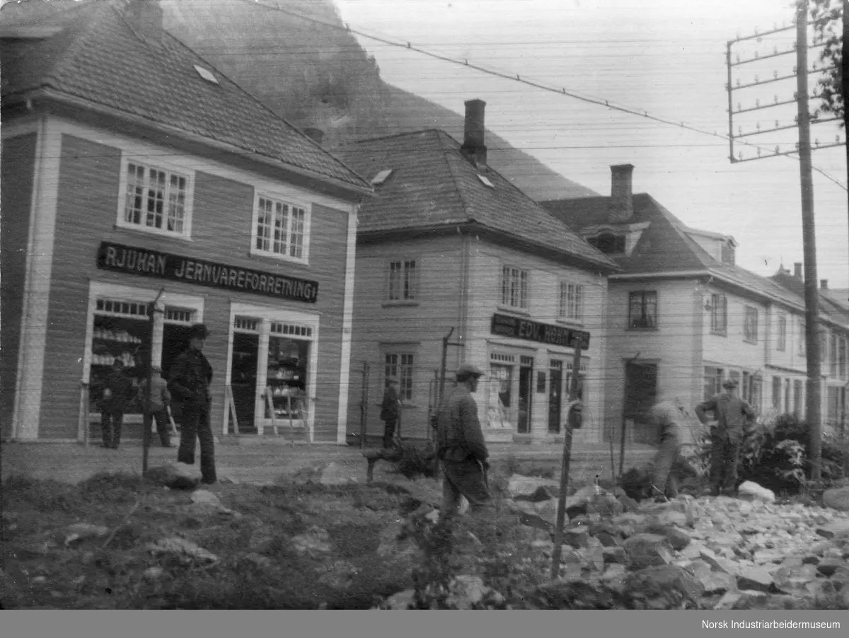 Arbeidskarer i gata utenfor butikkfasadene til Rjukan jernvareforretning og Edv. Køhn, i Sam Eydes gate.