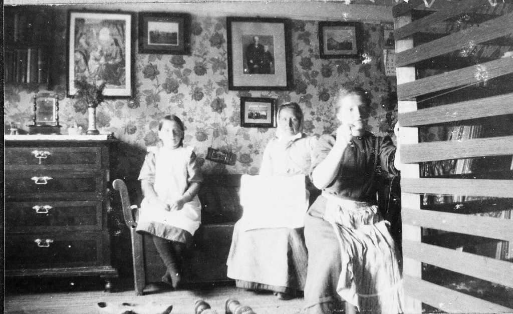 Interiør frå stova på Rimestad  med rennebom til høgre. Serina Rimestad (1857 - 1936) med døtrene Teresa (1898 - ) og Anna (1890 - ). Anna sit med rennebommen.