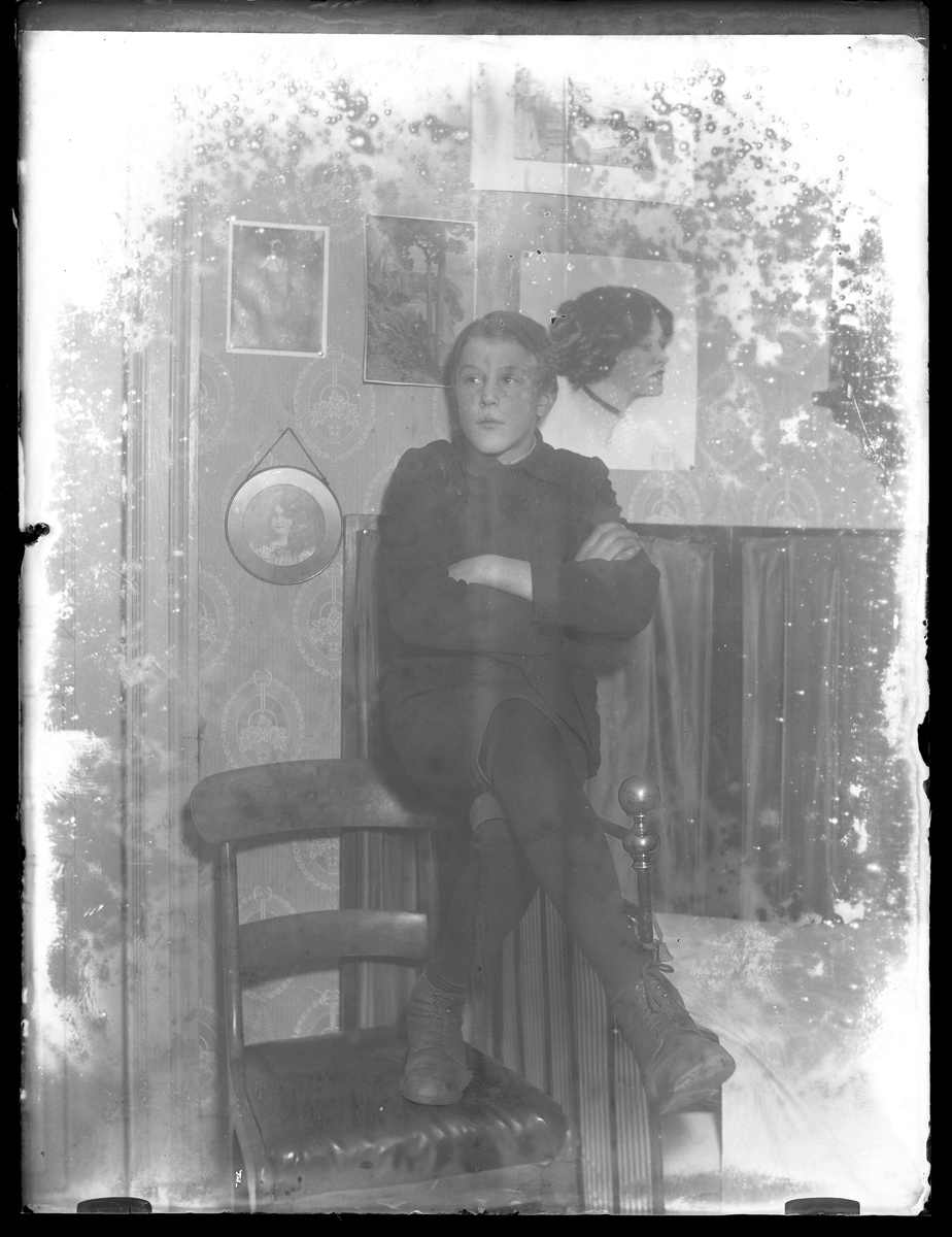 Arthur Andersson sitter med ben och armar i kors på en sänggavel av metall. På väggen bakom honom hänger tavlor med porträtt. Negativet är skadat.