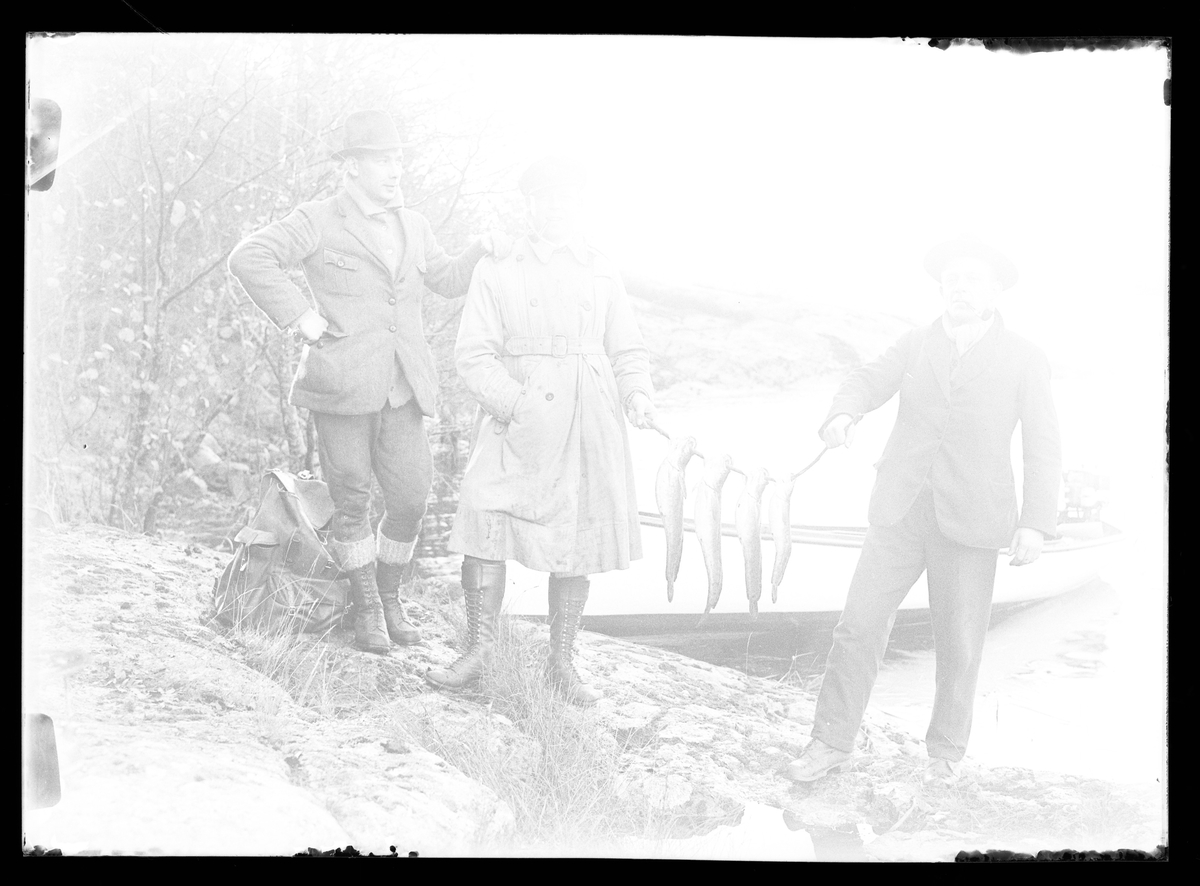 Överexponerat fotografi av Gustaf Claesson och Hjelm (möjligen John Hjelm) som håller upp fyra fiskar som de fångat. Bredvid står Harald Olsson och i bakgrunden syns en båt uppdragen på klipprona.
