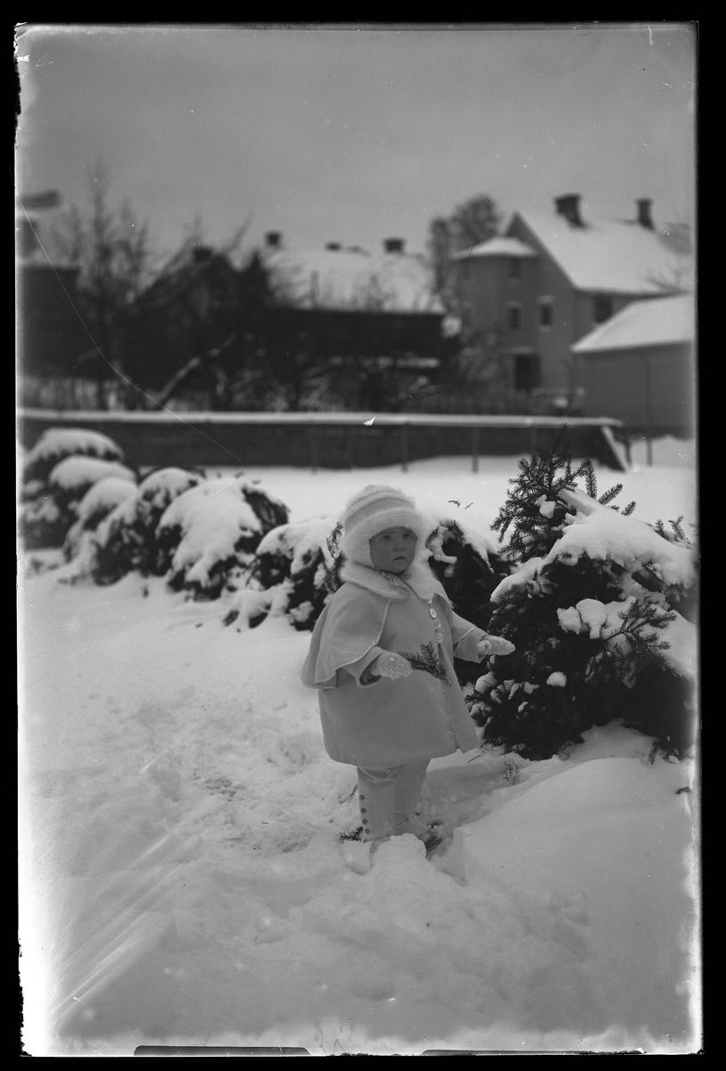 En liten flicka, tolkad som Elsa Margareta Berg, i vita vinterkläder står i snön med en kvist i handen. I bakgrunen syns ett lägenhetshus. I fotografens anteckningar står det "Ing[enjör] Bergs flicka".