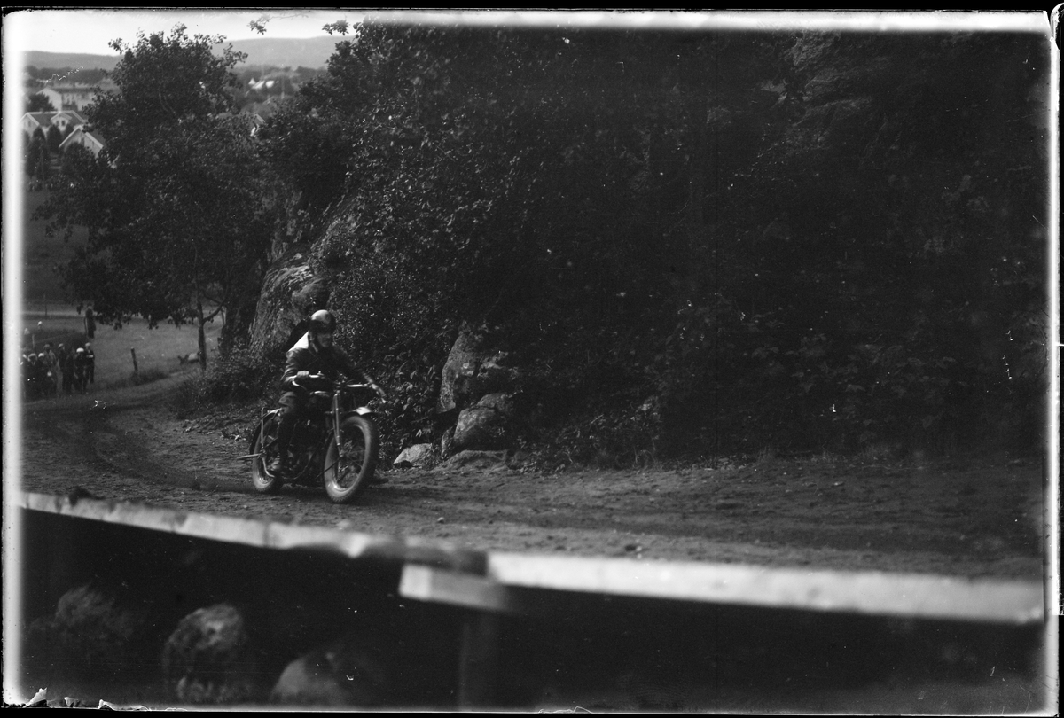 En person på motorcykel kör upp för en backe. I bakgrunden skymtar åskådare. I fotografens egna anteckningar står det "A.M.Ks [Alingsås motorklubb] backtävlan"