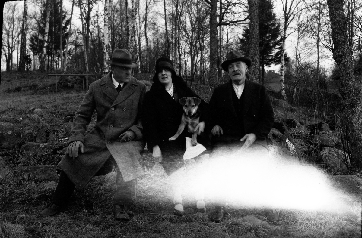 Lizzie Skoglund sitter mellan två män på en parkbänk. I famnen har hon en hund. I fotografens egna anteckningar står det "Skoglunds vid sjön".