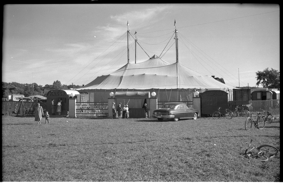 Ett rest cirkuställt med fyra tältstolpar.
Framför står en bil, cyklar står och ligger och runt om står olika vagnar.
En kvinna med ett barn i handen är på väg från tältet.

Tältet står på en stor gräsyta.