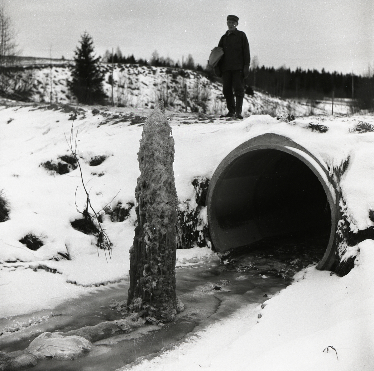 En man står på en bro och tittar ned på en ispelare i ett vattendrag den 19 december 1957.