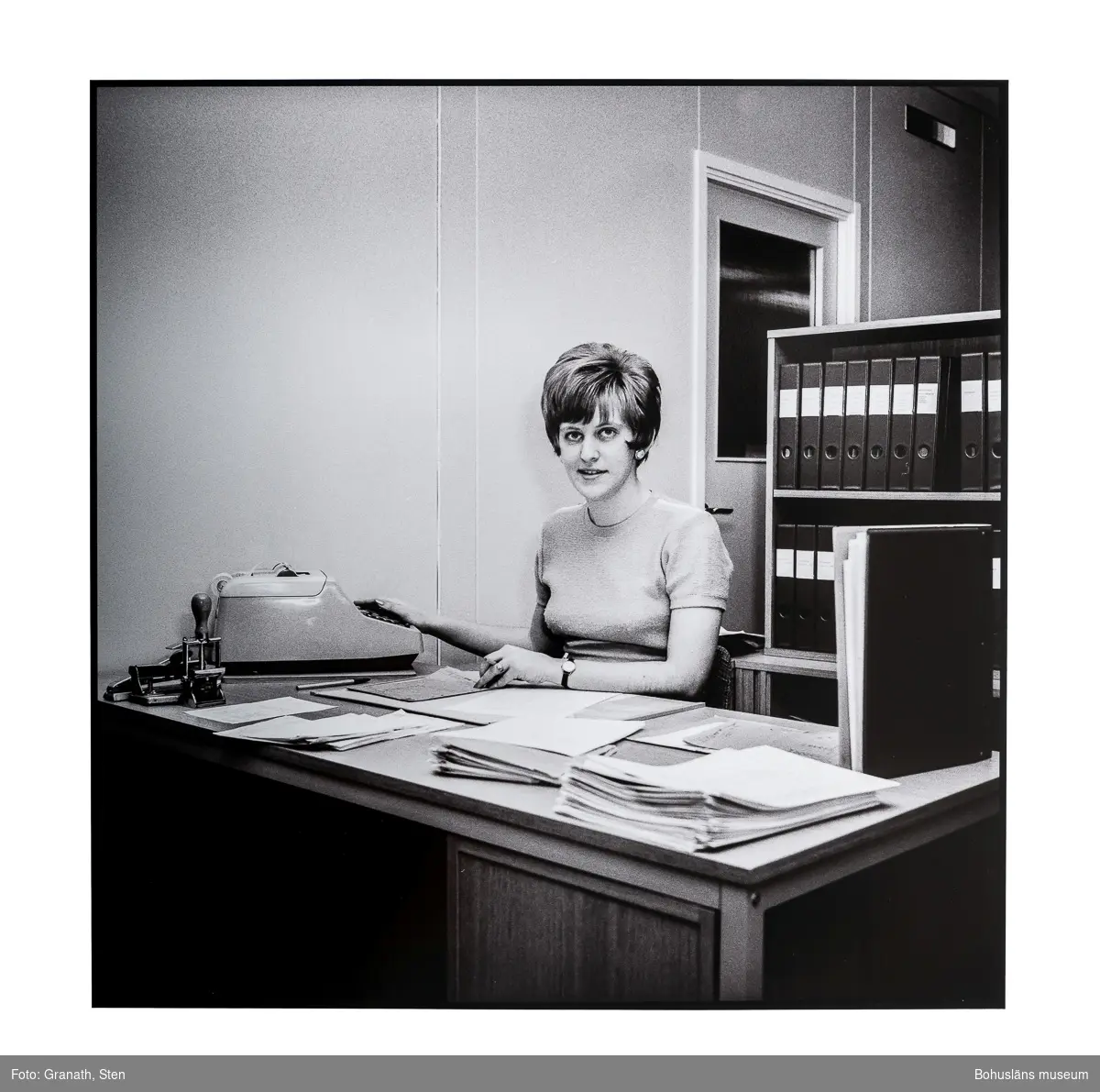 En kontorist på Domus sitter vid ett skrivbord och tittar mot betraktaren. En räknemaskin (eller skrivmaskin) står till höger på skrivbordet. Bakom henne står en bokhylla fylld med pärmar, denna verkar blockera en dörr i bakgrunden.