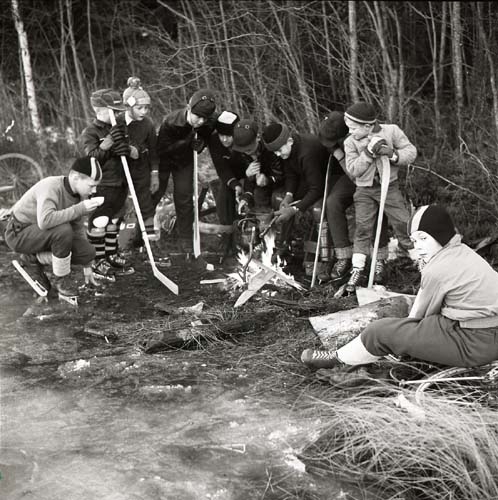 Glösbogänget på skridskor vid Stora Hisjön. En grupp ungdomar står runt en eld iförda skridskor och mössor. De håller också i  bandyklubbor,  27 december 1957.