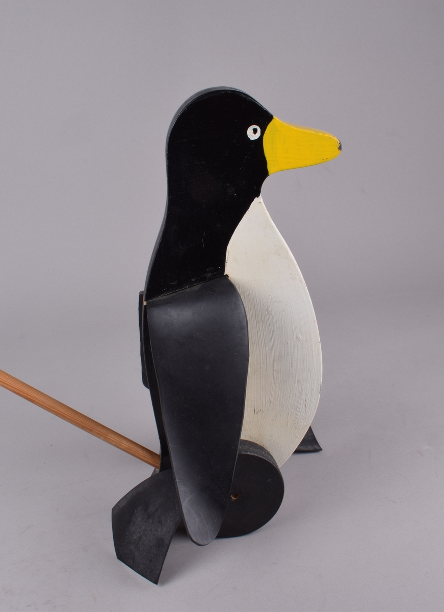 Utskåret pingvinfigur på to hjul med gummiføtter. Figuren er festet til en trepinne/håndtak. Når figuren skyves går føttene på hjulene rundt slik at pingvinen "går". En vinge av gummi festet på hver side.