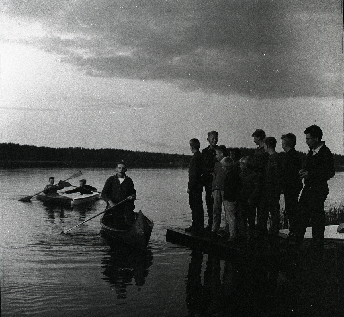 En grupp ungdomar nere vid en sjö. Några står på en brygga och andra paddlr kanot 1958.
