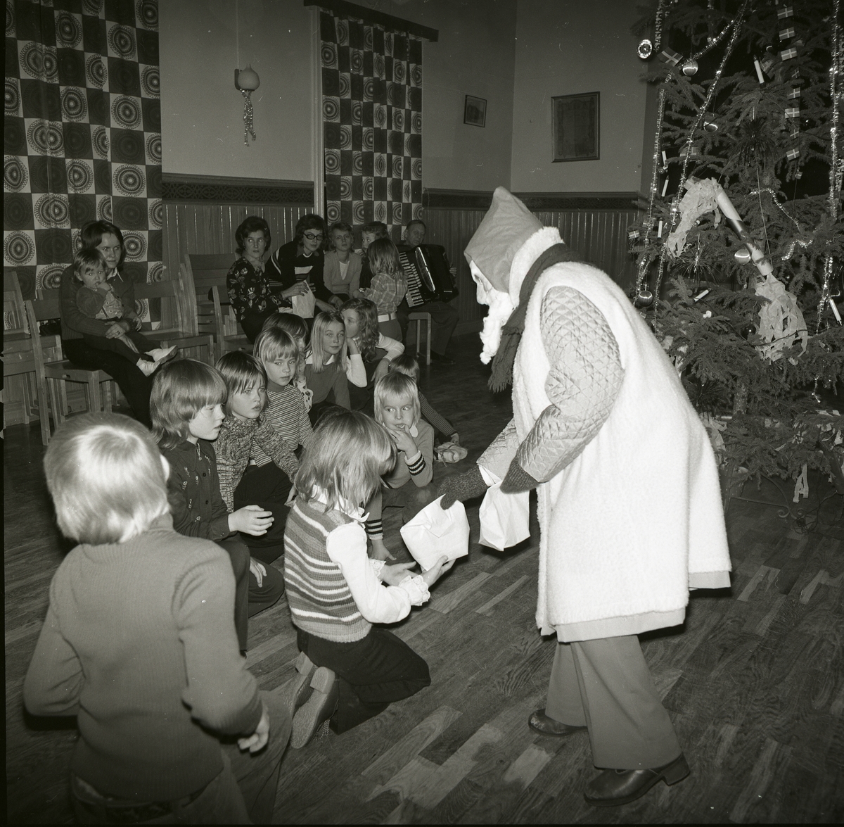 Julfest, januari 1974. Tomte delar ut påsar till förväntansfulla barn. I bakgrunden sitter en dragspelare och andra vuxna. Där står en gran klädd med flaggor och smällkarameller.