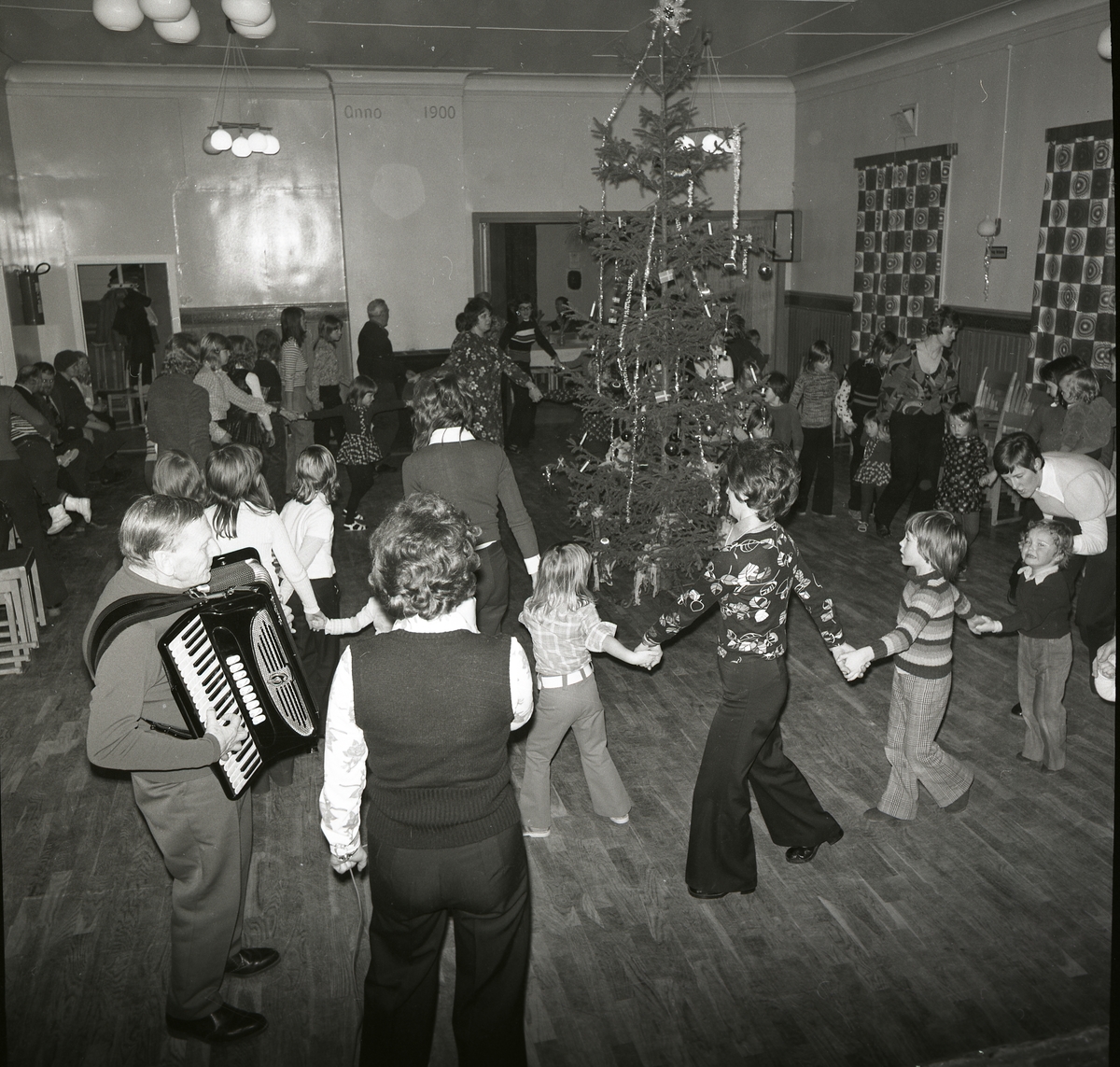 Julfest, januari 1974. En man spelar dragspel. Vuxna och barn dansar kring den klädda granen.