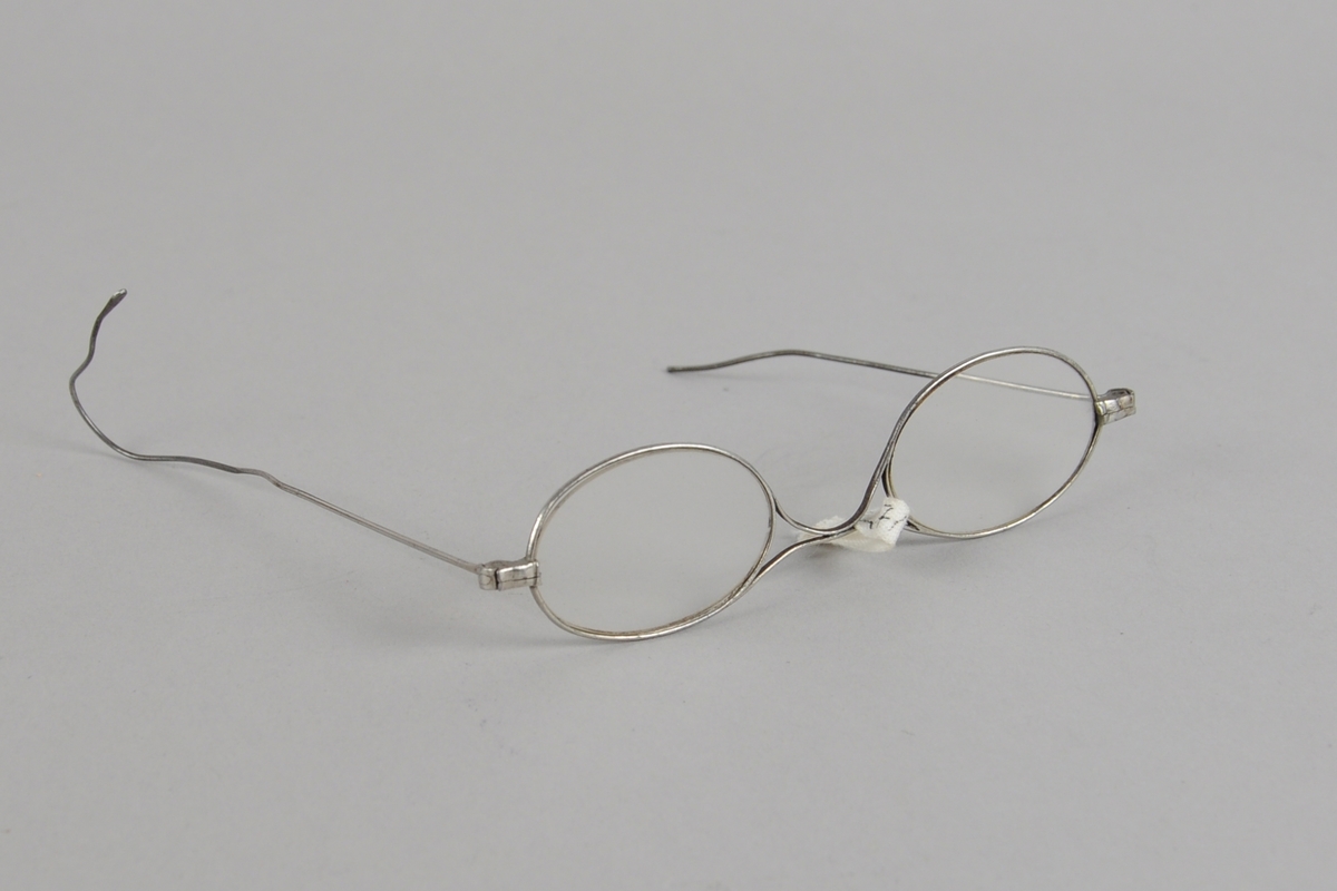 Ovale brilleglass med metallinnfatning. Den ene brillestangen er knekt, den andre er buet ytterst.