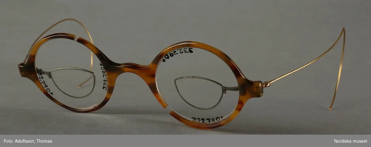 "Glasögon, frontstycket av celluoid, engelst nässtycke, spunna ridfjädrar av gulddoublé, glasen H. o V. cyl. -1,5 180° = sf. -0,5 D. runda 42 mm. P.D. 68 mm, bakom glasen små fällbara fattningar av vitmetall med biglas +2,5 D, glasögonen kunna användas som dubbelfokus, amerikanskt arbete från 1900-talets början." 
/Optikhistoriska museets katalog, nr. 272

Bågarna är trasiga på båda sidor, glasen sitter löst och kan ramla ut.
/Petrine Knight, 2015-08-13