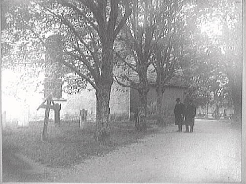 Två män i hatt och överrock står i en allé vid Slöinge kyrka. Invid kyrkan står gravvårdar av sten eller trä. På en utbyggnad till höger ligger ett sticktak.