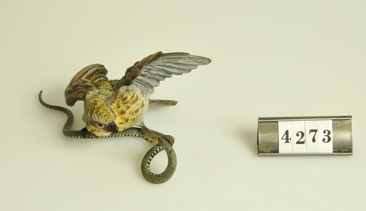 Gjuten metall målad i olika färger.
Figuren föreställer en fågel med en orm i klorna.
