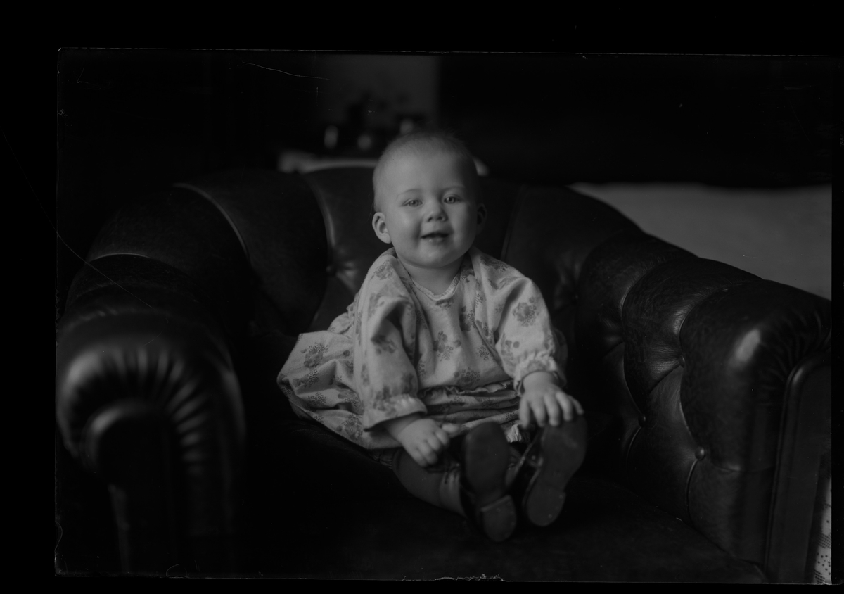 Ett litet barn i blommig klänning och kängor sitter i en chesterfieldfåtölj. I fotografens anteckningar står det "Nilssons barn (stadt)".