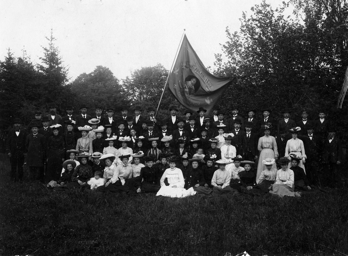 Gruppbild av ett 70-tal, stående och sittande, kvinnor, män, pojkar och flickor samt 2 småbarn. Männen är klädda i kostym, mössa och kubb (plommonstop). Kvinnorna i klänning och prydda hattar. En man längst bak i ledet håller en stor flagga med tydlig text "Gustav II Adolf". 
Templet Gustav II Adolf på sommarutflykt 1904. "Tagen den 5/6 kl. 10.00 1904"
