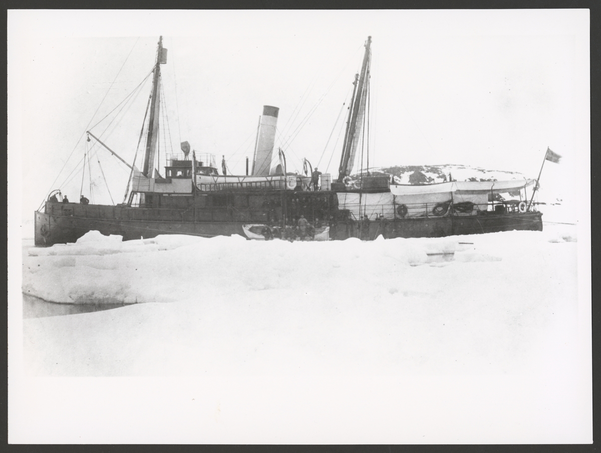Bilden visar kanonbåten Svensksund som ligger mellan stora isflak framför kusten av Spetsbergen från babordssida. En av räddningsbåtar ligger midskepps på isen. Manskapet är uppställt i båten och vid relingen för fotografering.