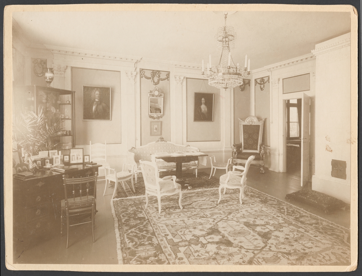 Bilden visar salongen (rum 114) i varvschefsbostället åren 1908-1912. Rummet är inred med klassicistisk interiör. Golvet är täckt med en stor matta och möblerna har arrangerats längs väggarna. Dörren står öppen och leder blicken vidare till salogen (rum 113).