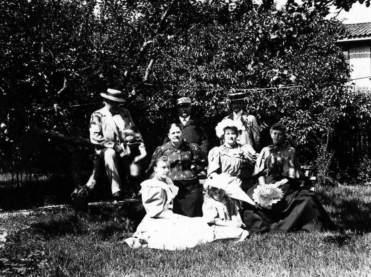 Gruppbild på tre män, fyra kvinnor och två barn som sitter i en trädgård, kvarteret Gustav.