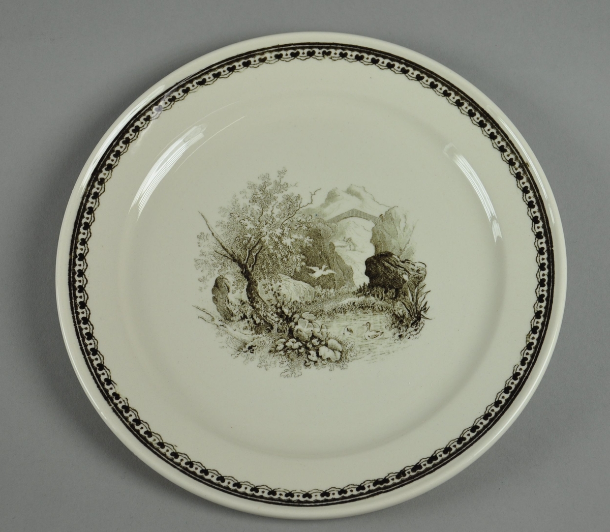 Asjett av glassert keramikk. Asjetten har svart dekortrykk av naturlandskap med bru i bakgrunnen, samt en borde langs randen.