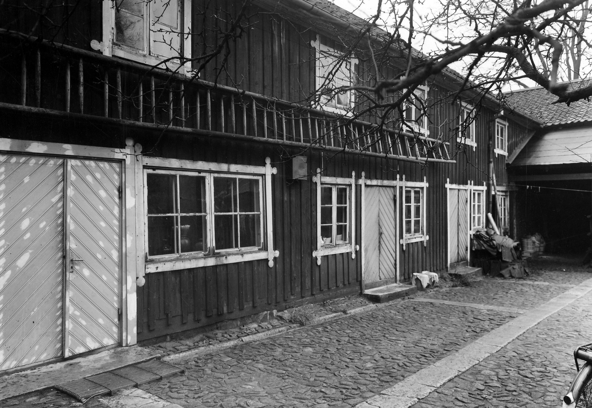Bilden visar tvåplansbyggnaden Apoteket Hjorten (1862-1946) från gårdspartiet med 3 separata ingångar. En lång stege är fäst på väggen mellan de två våningarna och gårdsplanen utanför är kullerstensbelagd.