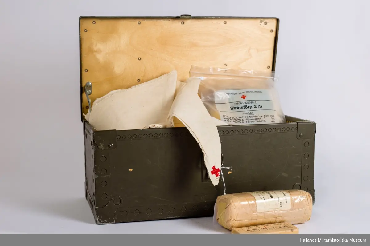 En låda i trä (41 x 16, 5 cm) med läderhandtag. Gröngrå färg med röda korsets emblem på. Lådan innehåller förbandspaket och diverse sjukvårdsmateriel. Vit klisterlapp ovanpå locket med röd text, handskrivet.