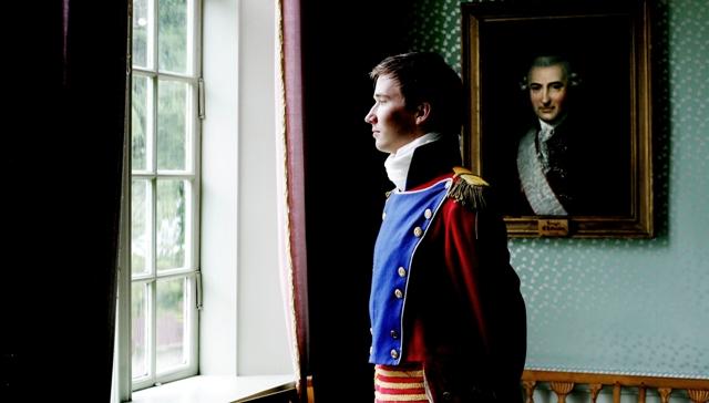 Ung mann i 1800-talls uniform står inne i Eidsvollsbygningen og ser tankefullt ut vinduet mot parken