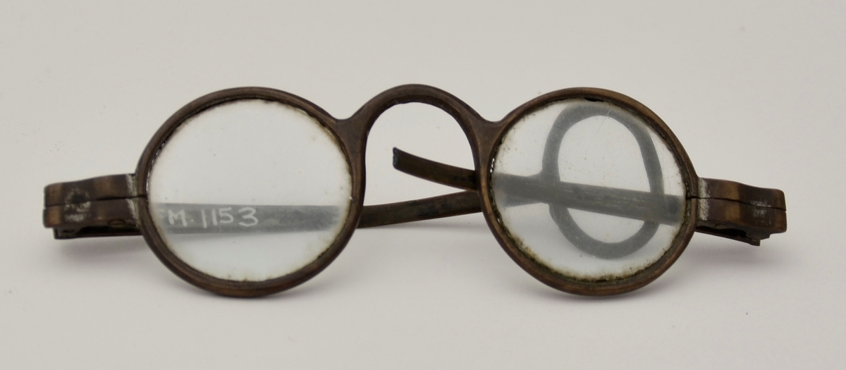 Briller. Sirkelrunde glas. Innfatning i brunt metall. Eine brillestang har avslutning med ring, den andre er kort, mogeleg knekt av.