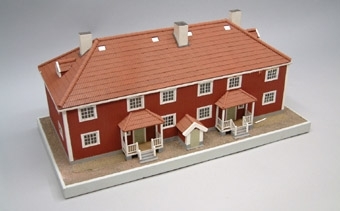 Modell i skala 1:50 av falurött tvåvåningshus med valmat sadeltak. Stående plank, tegeltak, och vita skorstenar. Byggnaden innehåller fyra lägenheter om 4 rum och kök. Inredning saknas.