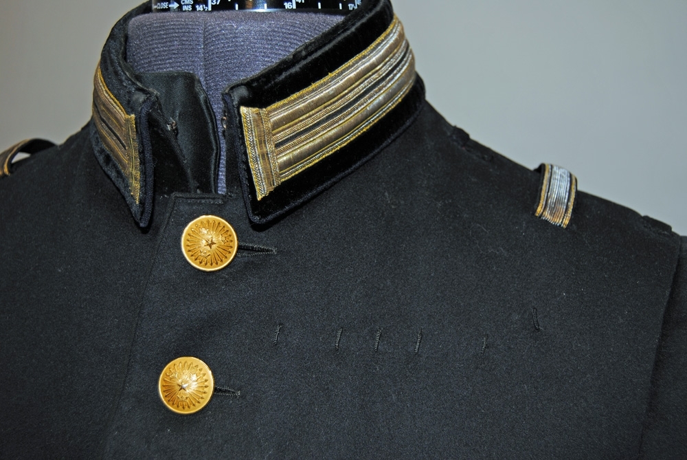 Vapenrock m/1886 av mörkblått kläde med svart sammet på krage och ärmuppslag. Rocken har avskärning i midjan och två formande insnitt fram. Den har svängd mitt-fram-linje, och knäpps med enradig knapprad bestående av 8 stycken (22 mm i diam) uniformsknappar av VVK:s egna: en femuddig stjärna omgiven av strålar på tre kronor. Fodrad med svart halvsiden i bålen, som är stickad i ett rutmönster i fram- och sidstycken.
Ärmarna är fodrade med smalrandigt vitt bomullstyg. Rocken har rå, dvs ofållade kanter nertill, och en 5 cm hög nedvikt krage av skjortkragsliknande modell. Kragen är mycket styv, troligen förstärkt inuti med papp eller kartong.
Rocken har två invändiga fickor - en stor påsydd ficka i vänster sida fram, samt en liten ficka med vertikal öppning strax till vänster om knappraden. Inne i rocken finns även ett svart midjeband med spänne.
Ärmuppslag och krage av svart sammet är prydda med två parallella 15 mm gyllene galoner som föreställer stiliserade knapphål. På ärmuppslagen sitter på varje sida två knappar i fusk-knapphål.
Det finns 6 sydda tränsar på rockens vänstra framsida för fastsättning av släpspännen eller ordnar, samt en sydd träns i vänster sida för att fästa bältet med sitt gehäng för värja.
Kragen stängs framtill med två stycken hyskar samt hakor. Inne i kragen finns två, ursprungligen tre, stycken tryckknappsliknande fästen, troligen för stärkkrage.
På axlarna är fastsytt på vardera sidan hällor för löstagbara axelklaffar samt epålettslejfar. Hällorna är av kläde medan epålettslejfen är klädd med enklare gyllene galon, 15 mm bred.
Rockens bakstycke är tredelat med både sidsöm och axelsöm liggande på ryggens baksida.
I skörtets baksida finns två formande insnitt på vardera sidan av mitt-bak-linjen, samt två vertikalt ställda fickor, så kallade fållklaffar) med tre knappar vardera. Fickpåsarna är av brunt bomullstyg.
Rockens foder är en satäng med varp av bomull eller lin och inslag av svart silke. Sammeten på krage och ärmuppslag är troligen en silkessammet.
Passpoal i svart kläde framtill från kragen ända ned, samt omkring ärmuppslag och på fållklaffar (bakfickor).