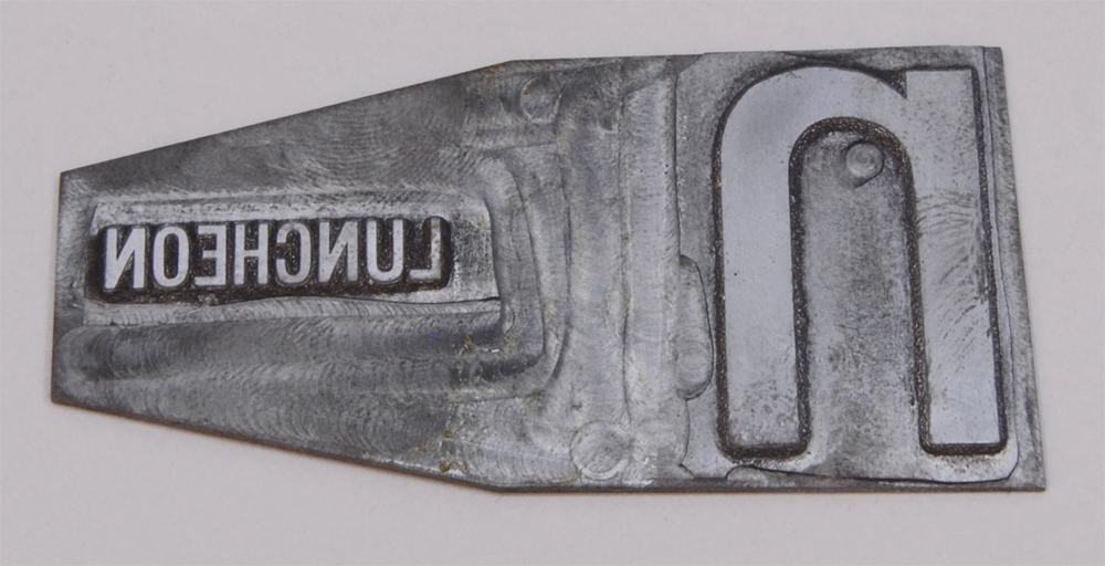 Rektangulär kliché av silverfärgad metall, de vänstra hörnen är beskurna. På höger sida finns bokstaven "n" i relief, och till vänster finns texten: "LUNCHEON", i relief och i ett mindre typsnitt.