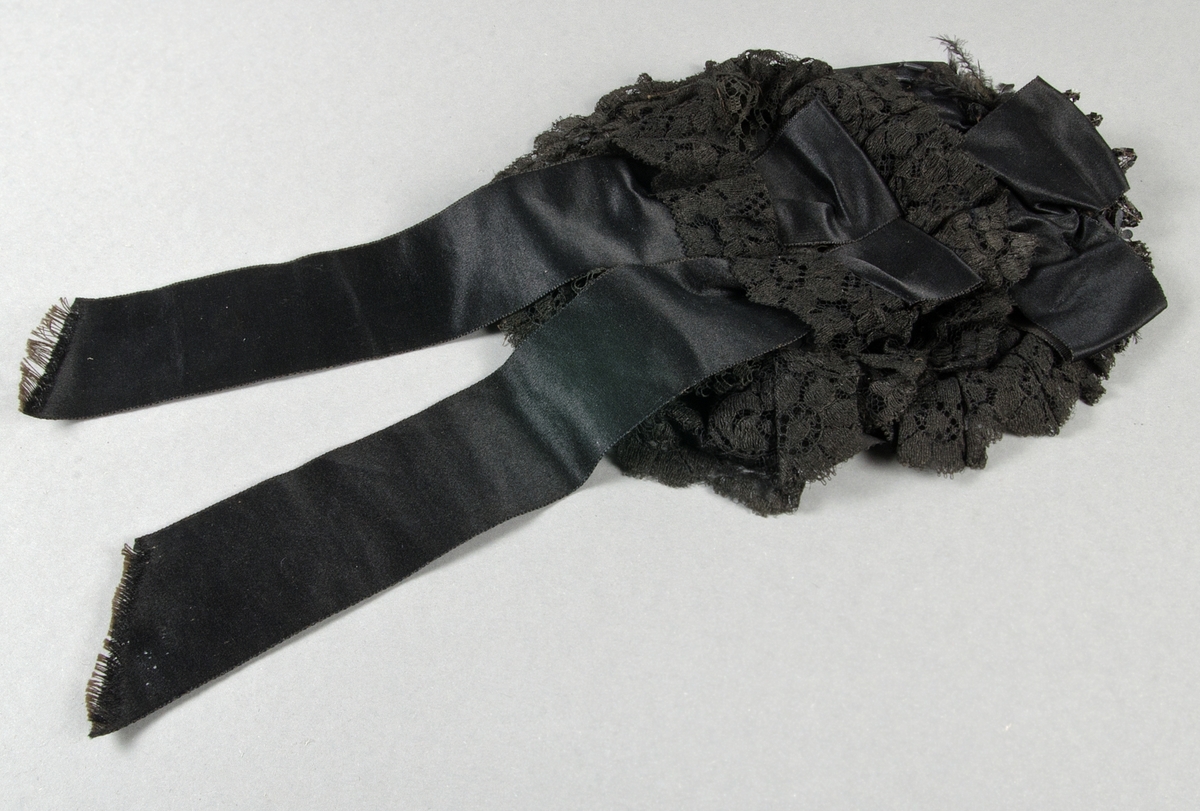 Hårklädsel av svart bomullspets med svarta duchesseband på oval ståltrådsstomme. Prydd med svarta pärlbroderier och fjädrar.

