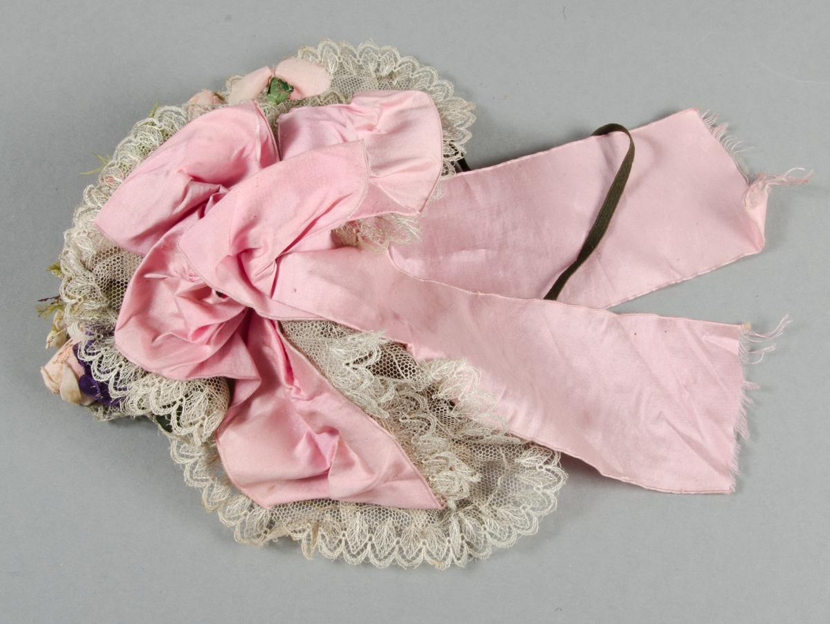 Hårklädsel, ungmorsmössa, av vit tyll med spetskanter och konstgjorda rosa, röda och blå blommor och gröna blad och fjädrar. Rosa sidenband i nacken. Nacksnodd av gummiband. Ståltrådsställning.