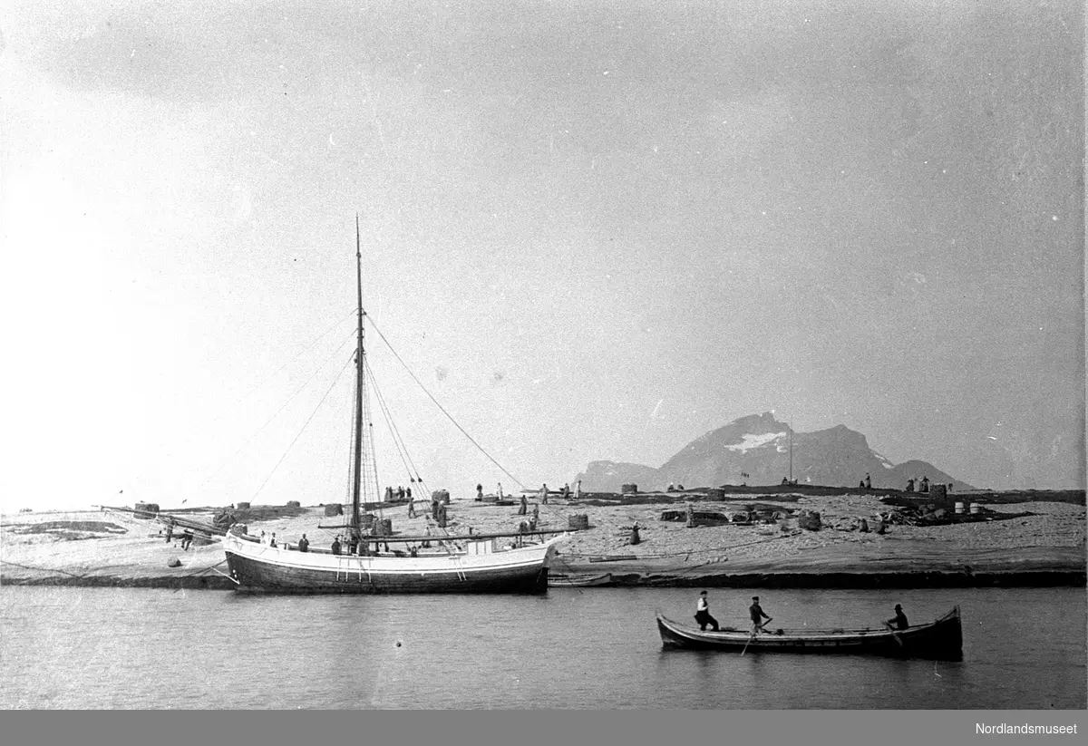 Gildeskål. Inndyr. Øya (?) Klippfiskberg med folk i arbeid. Klippfiskstabler. Ei jakt ligger foran, nordlandsbåt, tr. åttring ute på vågen. Fugløya bak. Tidlig i 1900-årene.