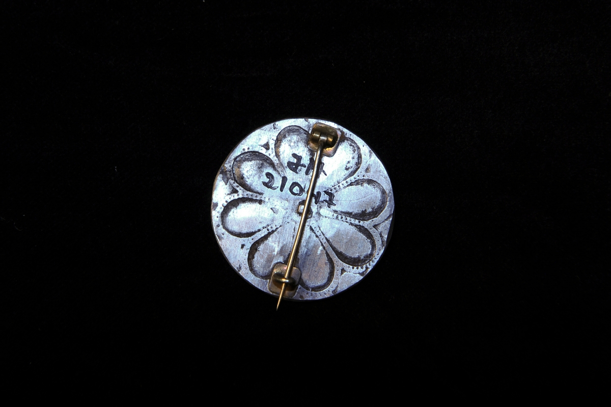 En rund brosch i förnicklat järn. Inpressat dekor i form av en blomma. Saknar märken eller stämplar.