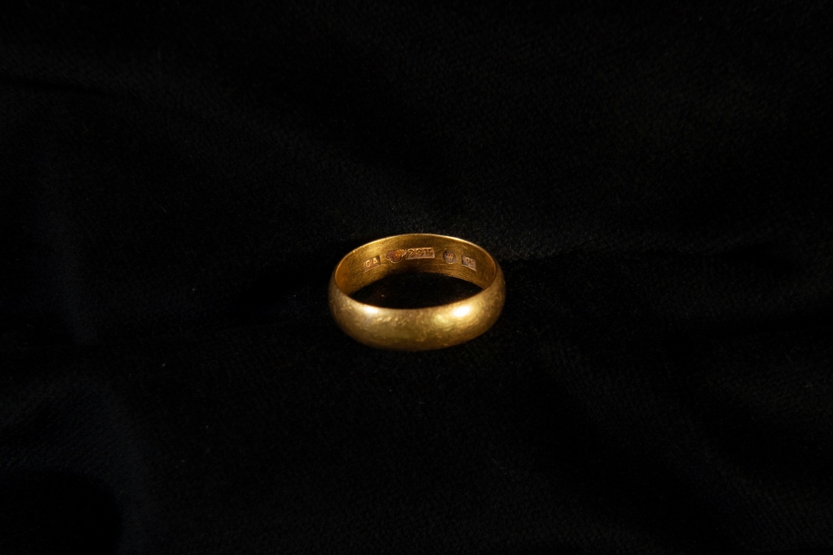 En vigselring av guld, bred, kupig. Förvaras tillsammans med JM.22794:1 i en röd pappask.