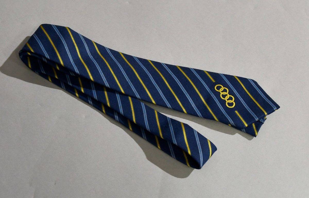 Mørkeblått slips med diagonale striper i gull og lyseblått. Nederst de olympiske ringene i gull. Slipset er designet for NK i Sverige.