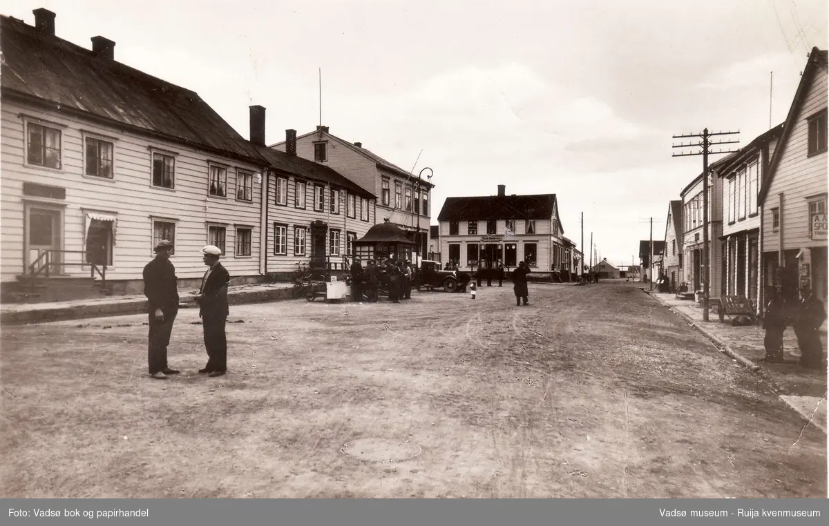 Tollbugata i Vadsø sett fra nord mot sør. Torget med en del folk, lastebil og kioskpaviljong. To menn i forgrunn. Mannen med hvit lue er Aksel Basma. Antatt 1930 tallet.