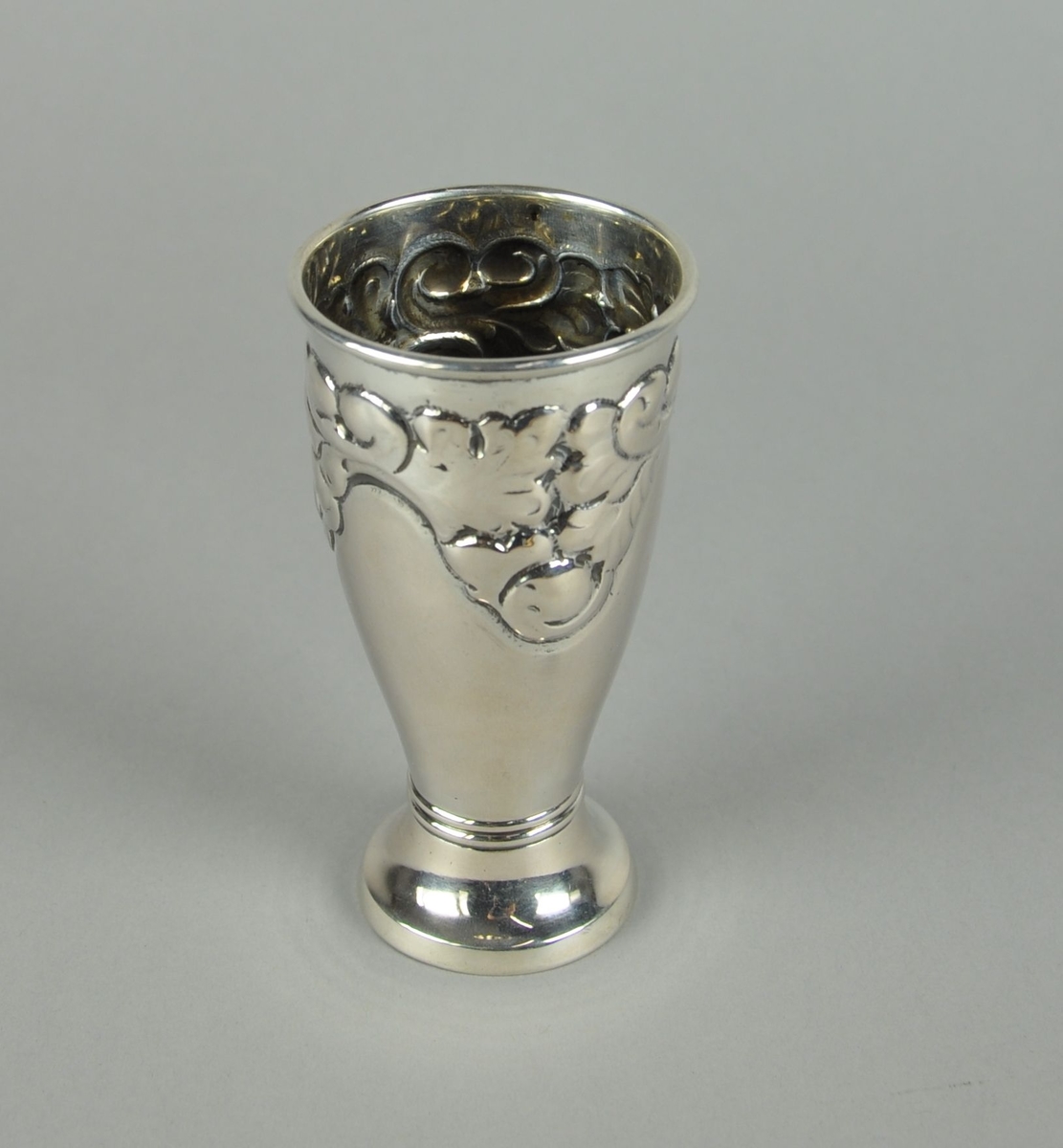 Pokal av sølv. Pokalen har sylindrisk form med innsnevret bunn. Utgående sokkel med to dekorative linjer. Øverst har pokalen akantuslignende gravert dekor.