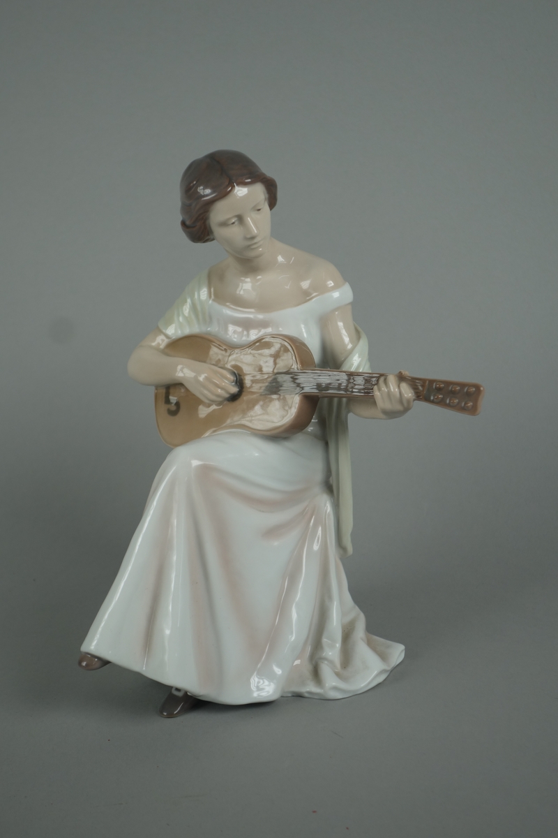 Porselensfigur formet som en gitarspillende dame iført lang hvir kjole og lysegult sjal. Hun sitter på en åttekantet tyrkisk taburett.
