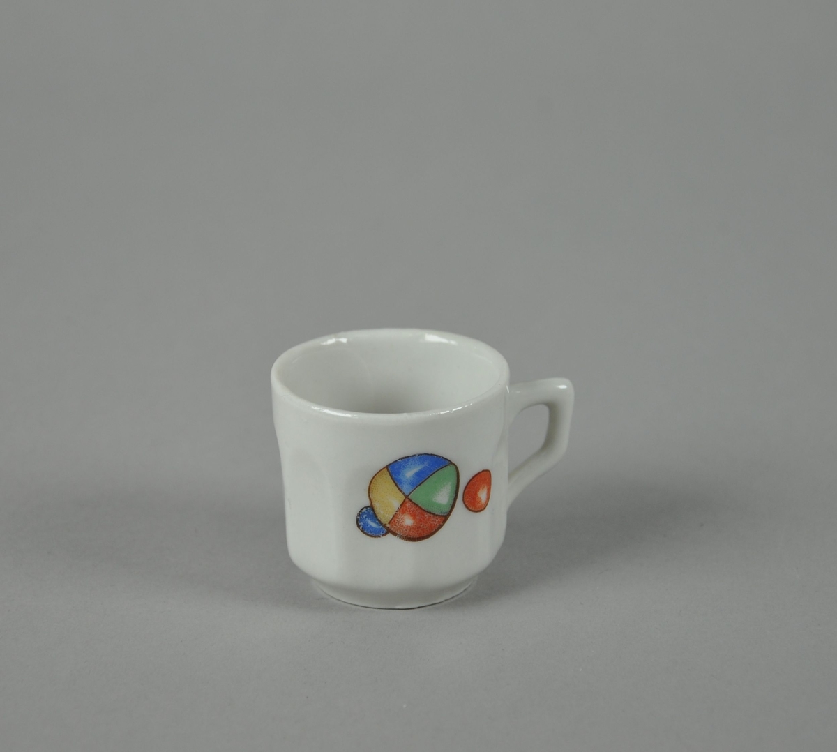 Kopp av glassert keramikk, med fasettert form. Hvit farge, med farget motiv av baller med forskjellige farger.