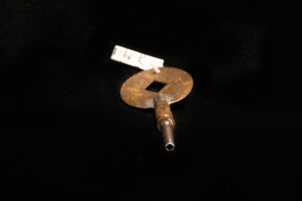 En urnyckel av mässing med runt handtag, inuti handtaget är ett snedställt kvadratiskt hål. Uppdragningsdelen är av metall. Det runda handtaget är gjort av en kinesisk penning.