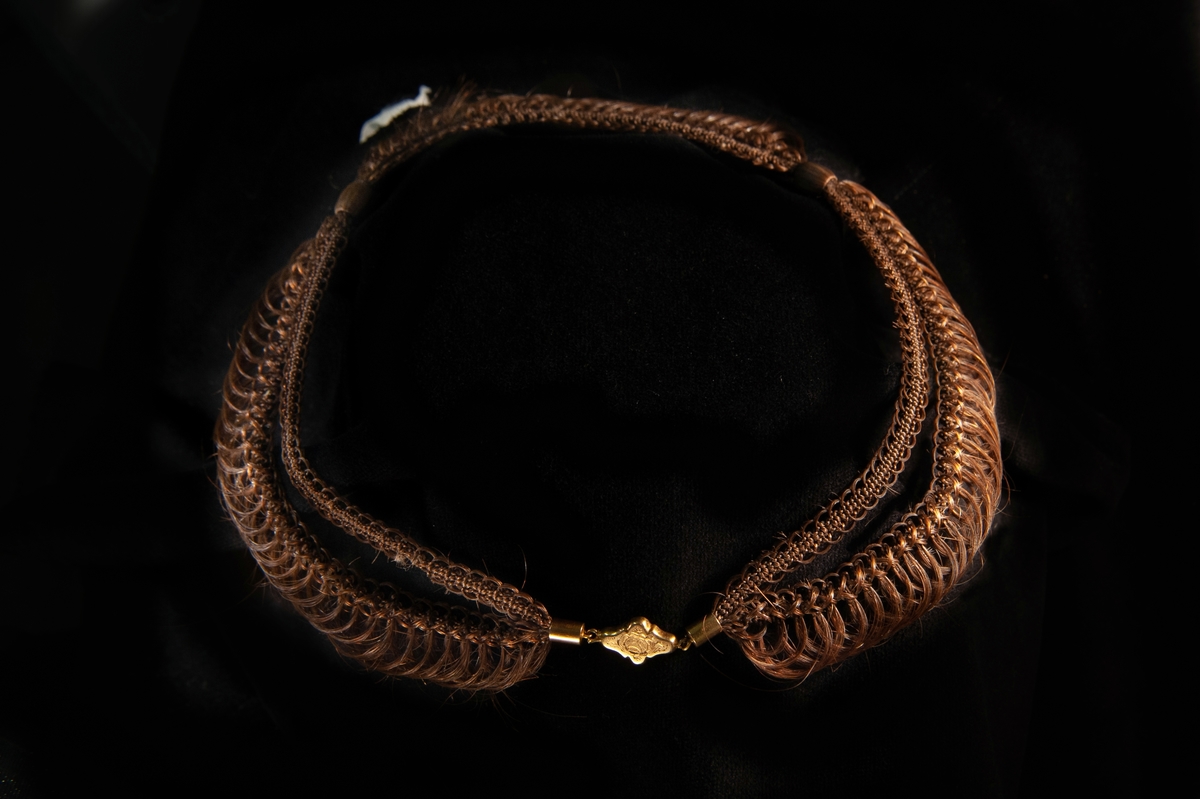 Ett dubbelflätat, tredelat halsband i mörkbrunt hårarbete. Sticklås i guld med graverad blomdekor på fronten. Stämplad på baksidan. 6 lösa hårtampar i mörkbrunt, längd: 180 - 240 mm.
