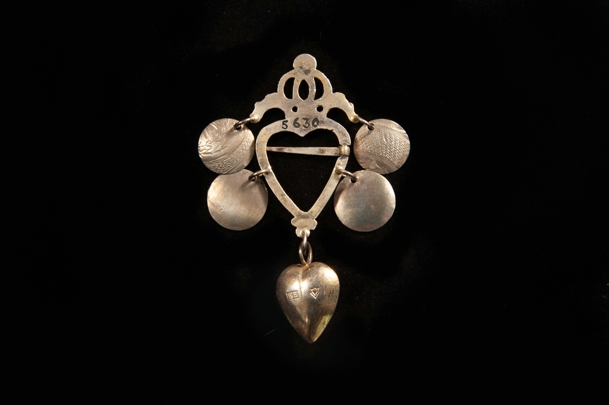 Hjärtformat spänne av förgyllt silver. Genombruten stiliserad krona upptill. Fyra skålar (varav de övre är graverade på ena sidan) hänger från spännet. Längst ner ett hjärta av silver (med annan stämpel: "E.E.., (Växjö) " (= Eric Ekström, Växjö, verksam cirka 1819-1840).